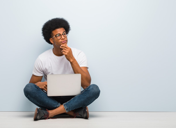homem de calça jeans e camisa branca sentado no chão de costas para uma parede branca e com um laptop no colo